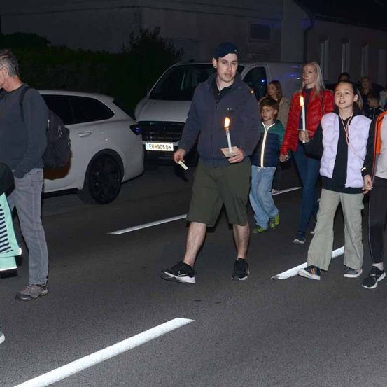 Eine Gruppe von Menschen läuft bei Nacht die Straße entlang und hält brennende Kerzen in den Händen. Ein Mann vorne hält eine brennende Kerze in einer Hand und eine Zigarette in der anderen. Am Straßenrand sind Autos geparkt.