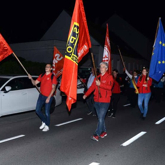 Eine Gruppe von Menschen marschiert mit Fahnen in der Hand auf der Straße. Sie tragen rote Jacken, Hosen und Turnschuhe.
