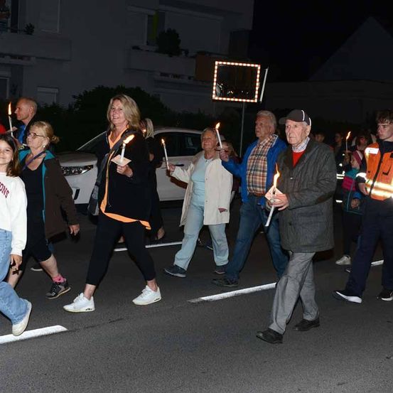 Gruppe von Menschen, die bei Nacht mit Kerzen in der Hand auf der Straße laufen. Eine Person in einer Warnweste läuft hinter ihnen.