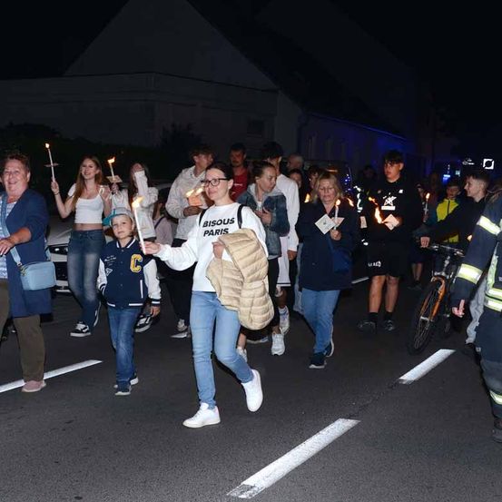 Menschen gehen bei Nacht mit Kerzen in der Hand und Jacken bekleidet die Straße entlang. Ein Kind hält eine Kerze und ein Mann vorne trägt einen Rucksack.