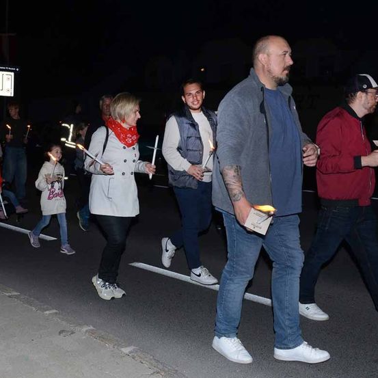 Eine Gruppe von Menschen geht bei Nacht auf der Straße und hält brennende Kerzen. Eine Frau im weißen Mantel und ein Mann im roten Mantel sind vorne.