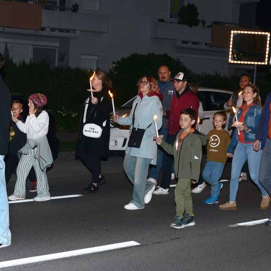 Eine Gruppe von Menschen zieht bei Nacht in einem Zug, hält Kerzen und trägt verschiedene Winterkleidung. Sie sind auf einer Straße, mit Gebäuden und einem Auto im Hintergrund.