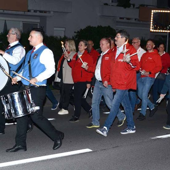 Eine Gruppe von Menschen in Rot und Blau zieht bei Nacht eine Straße entlang. Sie halten Kerzen, Trommeln und Stöcke.