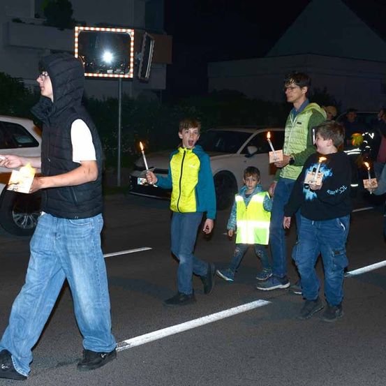 Eine Gruppe von Kindern und Erwachsenen geht bei Nacht mit Kerzen in der Hand auf einer Straße. Ein Mann in einer reflektierenden Weste führt sie an.