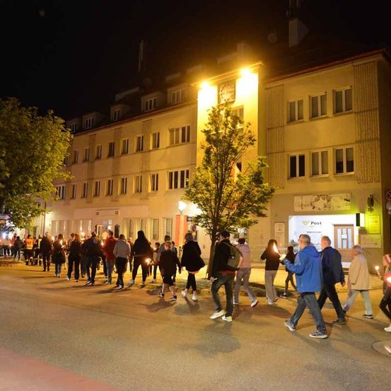 Eine Gruppe von Menschen läuft bei Nacht mit Kerzen in der Hand die Straße entlang. Die Straße wird von Straßenlaternen und beleuchteten Gebäuden erhellt.