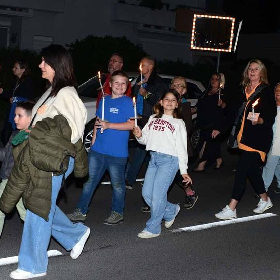 Mehrere Menschen, darunter Erwachsene und Kinder, gehen bei Nacht auf einer Straße. Sie tragen brennende Kerzen und nehmen anscheinend an einer Kerzenprozession teil.