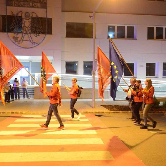 Gruppe von Menschen mit Flaggen, die auf einem Zebrastreifen vor einem Gebäude mit Logo und einer Person auf dem Bürgersteig gehen.