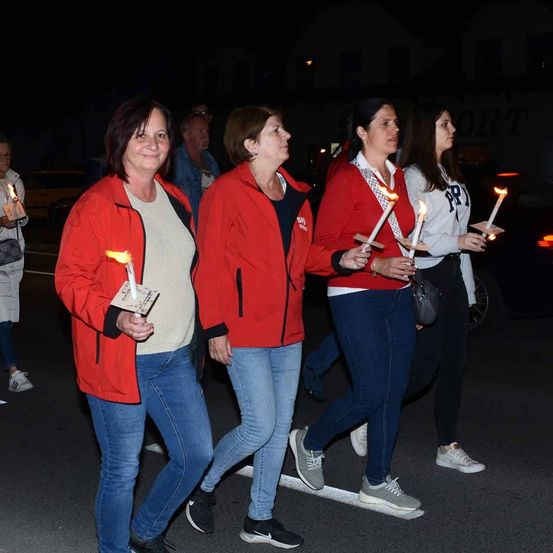 Fünf Frauen in roten Jacken laufen nachts auf der Straße. Sie halten brennende Kerzen in den Händen. Sie lächeln und nehmen anscheinend an einer Kerzenprozession teil.