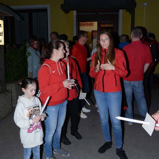 Menschen in roten Jacken stehen bei Nacht vor einem Gebäude und halten Kerzen. Eine Frau spricht. Hinter ihnen steht eine Gruppe von Menschen in roten Hemden, und ein gelbes Schild ist an der Wand zu sehen.