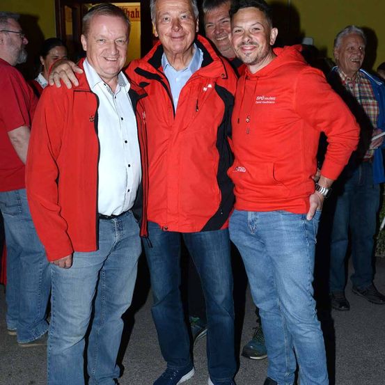 Drei Männer, einer in einem roten Jacke und zwei in weißen Hemden, stehen vor einer gelben Wand mit anderen in roten und karierten Hemden.