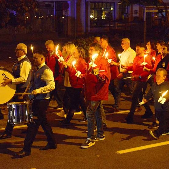 Eine Gruppe von Menschen in roten Jacken mit brennenden Kerzen, die nachts auf der Straße marschieren.