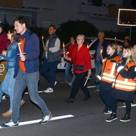 Eine Gruppe von Menschen, darunter Erwachsene und Kinder, halten brennende Kerzen in der Hand und gehen auf einer Straße.