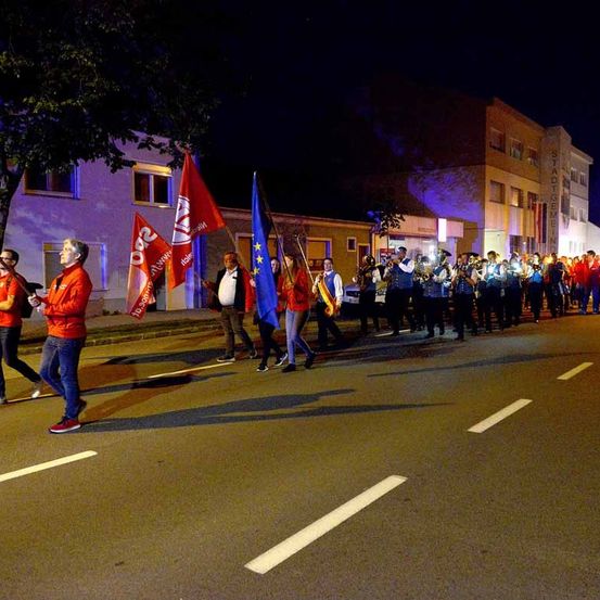 Eine Gruppe von Menschen marschiert nachts die Straße entlang, einige schwenken Flaggen und andere spielen Musikinstrumente.