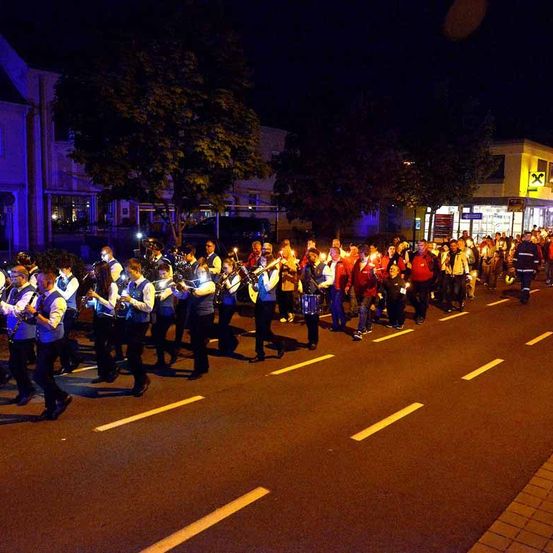 Eine Gruppe von Menschen marschiert bei Nacht auf der Straße. Sie spielen Musikinstrumente wie Trompeten, Trommeln und Saxophone. Einige von ihnen halten Kerzen.