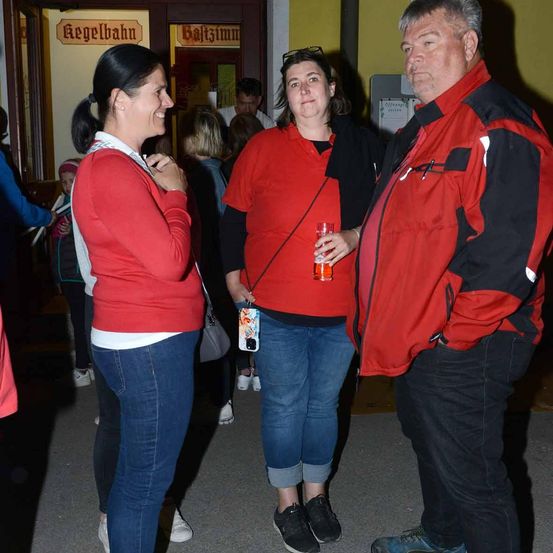 Zwei Frauen und ein Mann in roten Jacken stehen in einem Innenraum, wahrscheinlich einem Bahnhof, und führen ein Gespräch.