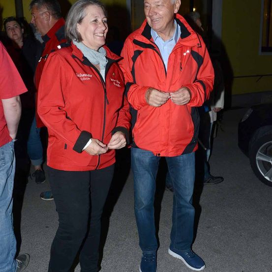 Ein älteres Paar in roten Jacken steht nachts draußen, lächelt und schaut auf etwas. Sie sind von anderen Leuten in Jacken umgeben, von denen einer einen Hut trägt.