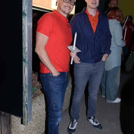 Zwei Männer, einer in einem roten Shirt und der andere in einer blauen Jacke, stehen nebeneinander. Sie halten beide eine Kerze. Hinter ihnen hält eine weitere Person in einer weißen Jacke ebenfalls eine Kerze.