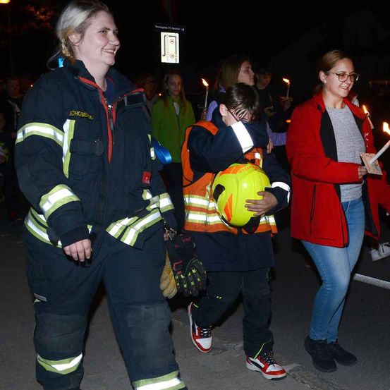 Drei Menschen gehen im Dunkeln zusammen. Eine Frau in einer Feuerwehruniform, eine Frau in einer roten Jacke und ein Junge mit einem gelben Helm.