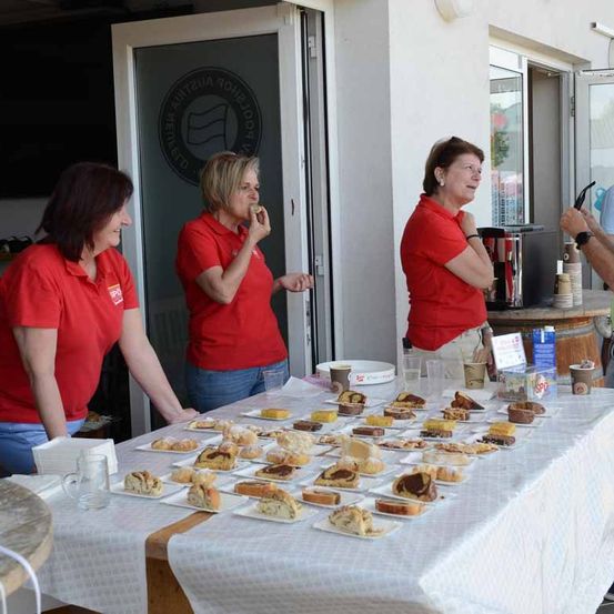 Drei Frauen in roten Shirts stehen um einen Tisch voller Desserts. Die Frau auf der rechten Seite hält eine Tasse in der Hand.