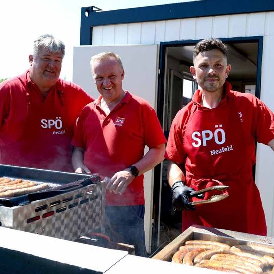 Drei Männer in roten T-Shirts mit dem Text 'SPO Neufeld' stehen vor einem Container. Zwei grillen Würstchen und einer hält eine Zange.