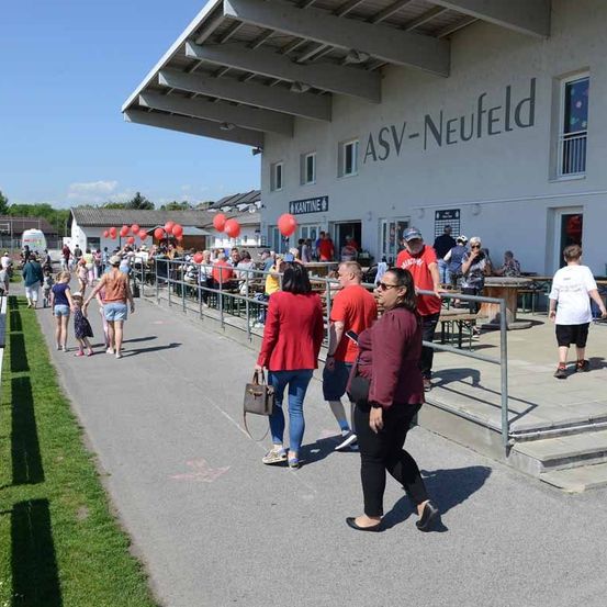 Eine Menschenmenge geht auf einem gepflasterten Bereich vor dem ASV-Neufeld-Gebäude entlang. Einige Leute tragen Sonnenbrillen, Hüte und Turnschuhe. Es gibt Tische und Bänke an der Seite des Gebäudes.