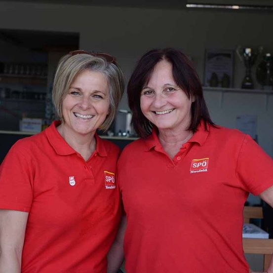 Zwei Frauen lächeln und posieren für ein Foto. Sie tragen rote Poloshirts mit dem Logo von SPO Neufeld. Hinter ihnen steht ein Regal mit einigen Trophäen.
