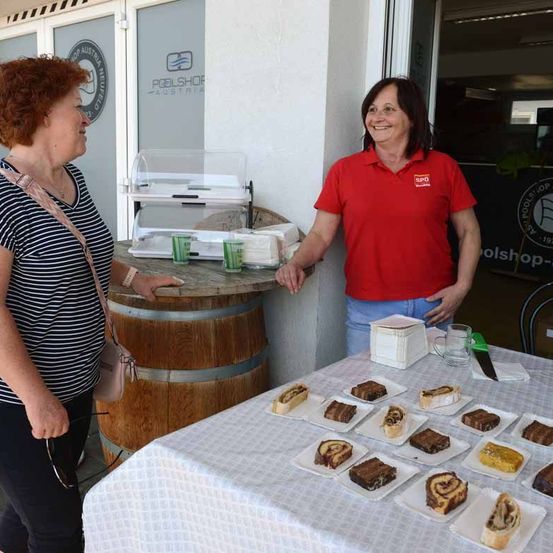 Zwei Frauen stehen vor einem Tisch mit Desserts, einem Holzfass und einer Glastür. Die Frau rechts trägt ein rotes Shirt und lächelt.