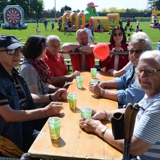 Eine Gruppe älterer Erwachsener sitzt an einem Holztisch und trinkt aus grünen Bechern. Der Tisch ist von einem Metallzaun umgeben. Dahinter steht eine Frau und hält einen Ballon. In der Ferne spielen Menschen und es gibt aufblasbare Strukturen.