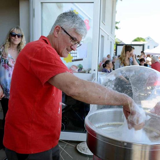 Ein Mann in einem roten T-Shirt macht Zuckerwatte in einer Maschine. Eine Frau mit Sonnenbrille und Blumenmuster steht hinter ihm, und mehrere Personen sind im Hintergrund versammelt.