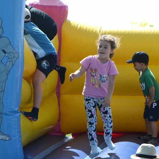 Drei Kinder spielen in einem großen aufblasbaren Hüpfhaus. Ein Kind klettert an der Seite, während ein anderes Kind davor steht. Das dritte Kind trägt eine Mütze und Turnschuhe.