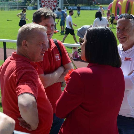 Vier Erwachsene stehen bei einer Outdoor-Veranstaltung zusammen; drei Männer in roten T-Shirts und eine Frau in einem roten Blazer. Im Hintergrund, in Unschärfe, sind Menschen bei verschiedenen Aktivitäten auf einem Rasenfeld zu sehen.