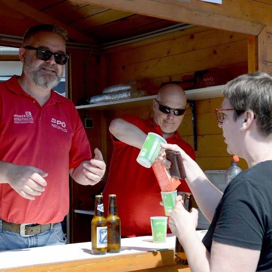 Drei Menschen sind in einem hölzernen Bar-Stand, zwei Männer tragen rote Polo-Shirts und eine Frau trägt ein schwarzes Shirt. Der Mann vorne trägt eine Sonnenbrille und gibt einen Daumen hoch.