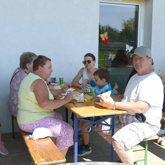 Eine vierköpfige Familie sitzt an einem Holztisch und isst draußen vor einem Gebäude mit Glastür.