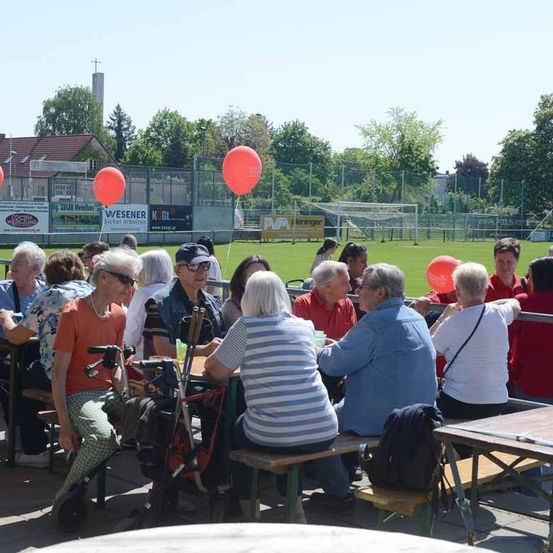 Eine Gruppe älterer Menschen sitzt auf Bänken in einem Außenbereich. Einige von ihnen tragen Sonnenbrillen. Es gibt zwei rote Luftballons am Zaun. In der Ferne befindet sich ein Fußballfeld.