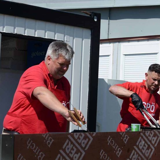 Zwei Männer in roten Hemden und Schürzen kochen vor einem Gebäude. Ein Mann benutzt Zangen und der andere hält einen Becher. Hinter ihnen steht ein Lagerkasten. Die Wand hat eine weiße Klappe und ein Logo. Der Boden ist mit einem Holzboden bedeckt.