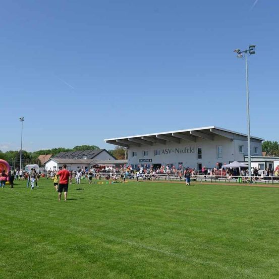 Eine Gruppe von Menschen versammelt sich auf einem Sportplatz, wahrscheinlich für eine Sportveranstaltung. Im Hintergrund steht ein großes Gebäude mit der Aufschrift ASV-Neufeld. Straßenlaternen und eine aufblasbare Burg sind ebenfalls zu sehen.