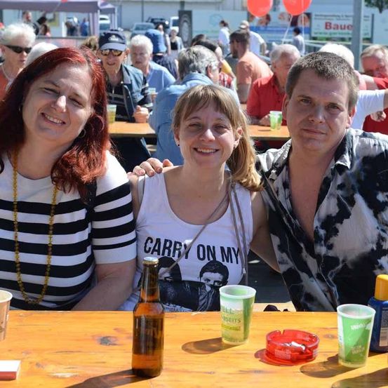 Drei Menschen lächeln in die Kamera, zwei Frauen und ein Mann. Sie sitzen an einem Tisch mit Gläsern und einer Bierflasche. Hinter ihnen sitzen weitere Menschen an Tischen, und es gibt einige Luftballons und ein Schild.
