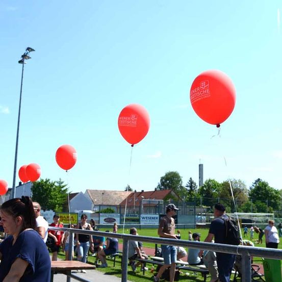 Rote Luftballons mit dem Text Wiener Stadtische schweben in der Luft über einem Sportplatz, wo mehrere Menschen auf Bänken sitzen und die Veranstaltung beobachten.
