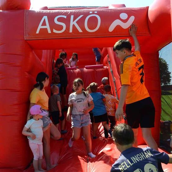 Eine Gruppe von Kindern und Erwachsenen spielt in einem großen aufblasbaren Hindernisparcours mit dem Wort 'ASKO' oben.