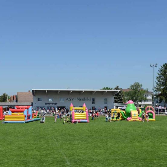 Eine Gruppe von Kindern spielt in aufblasbaren Spielgeräten auf einem Rasenfeld. Das Feld befindet sich in der Nähe eines Gebäudes mit der Aufschrift ASV-Neufeld. In der Ferne stehen Bäume und Straßenlaternen.