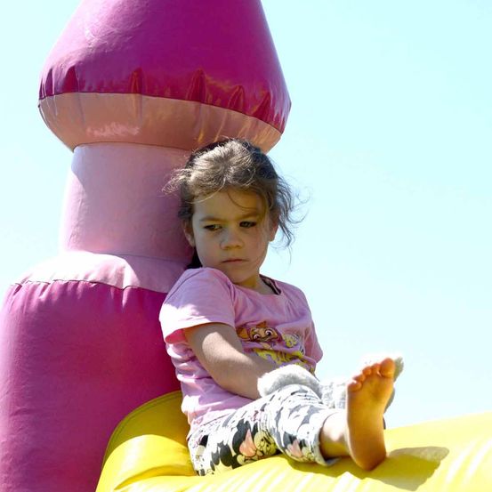 Ein kleines Mädchen mit braunen Haaren sitzt auf einem gelben aufblasbaren Schlitten mit den Füßen auf dem rosa aufblasbaren Schloss.