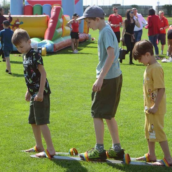 Drei Jungen balancieren auf einem Seil auf einem grünen Feld. Einer von ihnen trägt bunte Sandalen. Im Hintergrund steht ein aufblasbarer Hüpfburg und mehrere Personen.