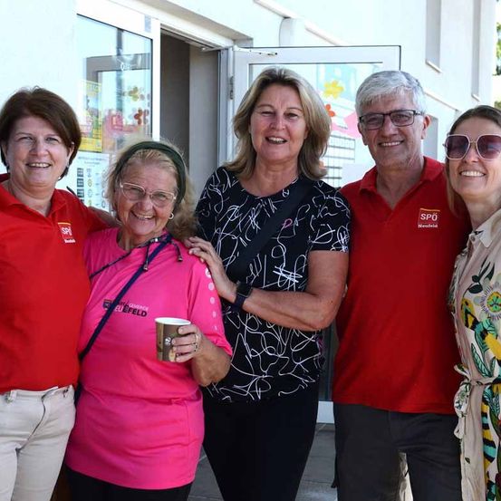 Fünf Erwachsene lächeln in die Kamera, zwei Frauen in roten Shirts, eine mit einer Tasse, eine Frau in einem schwarz-weißen Shirt und ein Mann in einem roten Shirt.