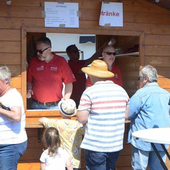 Eine Gruppe von Menschen steht vor einem hölzernen Verkaufsstand. Im Inneren des Standes steht ein Mann hinter der Theke, und ein anderer Mann hält ein weißes Schild mit dem Wort 'Etrank'. Es befinden sich zwei Personen im Stand, von denen einer einen Hut trägt.