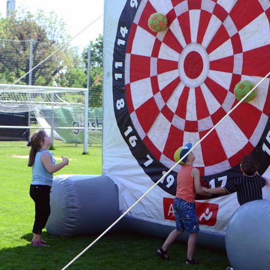 Drei Kinder spielen mit einem riesigen aufblasbaren Dartboard. Das Mädchen steht vor dem Dartboard, während zwei Jungen den Dart halten und versuchen, ihn zu werfen.