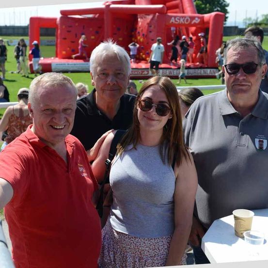 Drei Menschen stehen und lächeln für ein Selfie. Eine Frau in der Mitte hat einen Rucksack. Sie befinden sich in einem Außenbereich mit einer Tasse auf einem Tisch. Im Hintergrund sind mehrere Personen zu sehen, und dahinter befindet sich ein aufblasbares Spielplatz.