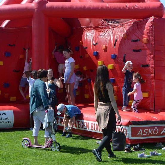 Mehrere Kinder und Erwachsene spielen auf einer riesigen roten aufblasbaren Wand mit verschiedenfarbigen Quadraten. Einige klettern, während andere auf dem Boden stehen. Eine Frau geht vor der aufblasbaren Wand und hält eine Handtasche.