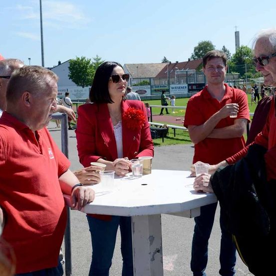 Eine Gruppe von Menschen steht um einen weißen Tisch im Freien. Eine Frau in der Mitte hält eine rote Blume. Der Mann rechts trägt eine Sonnenbrille und einen Becher. Der Mann links trägt ein Armband. Es gibt Becher auf dem Tisch.