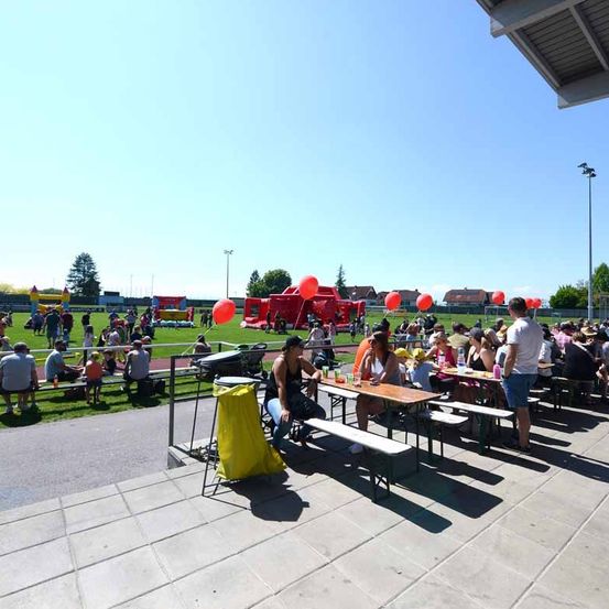 Eine Gruppe von Menschen sitzt auf Bänken und isst, während einige Leute vor den Tischen stehen. In der Ferne befinden sich aufblasbare Objekte und im Bereich sind Straßenlaternen.