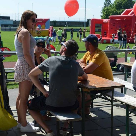 Menschen sitzen an einer Picknick-Tisch im Freien. Eine Frau steht neben dem Tisch und schaut auf die sitzenden Personen. Im Hintergrund sind andere Leute, die herumlaufen und spielen.